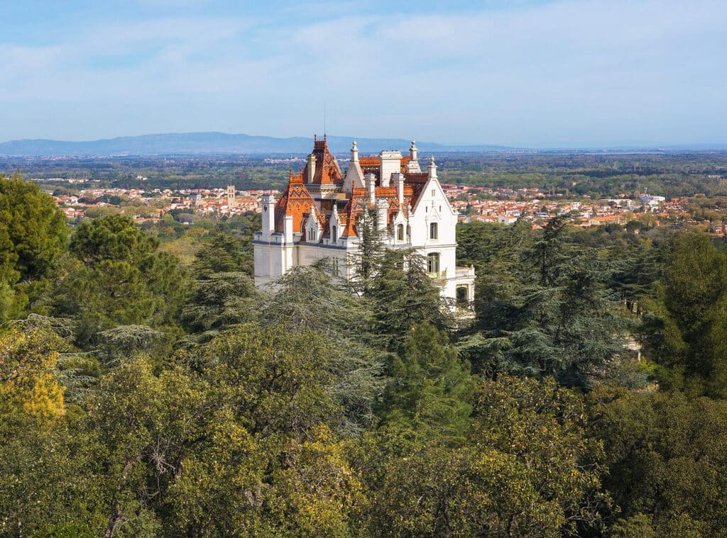 Château blanc aux toits rouges parmi arbres et montagnes.