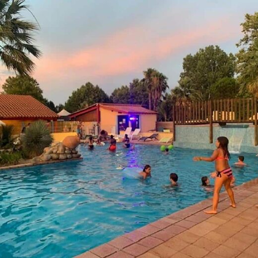 Vacanciers profitant de la piscine au camping Arlequin Vacances, palmiers et animations au coucher du soleil.